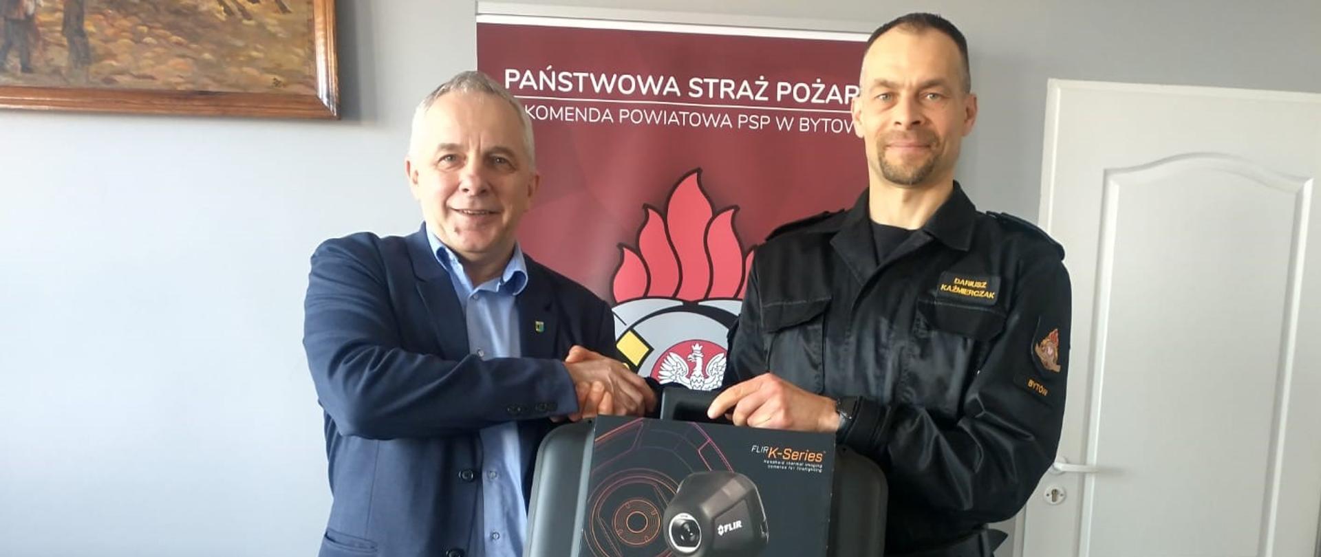 Frontem do zdjęcia stoją Komendant Powiatowy PSP w Bytowie oraz Starosta Bytowski. Podają sobie ręce. Komendant trzyma walizkę z kamerą termowizyjną. W tle widać czerwony plakat z napisem Państwowa Straż Pożarna - Komenda Powiatowa PSP w Bytowie oraz logiem straży pożarnej.