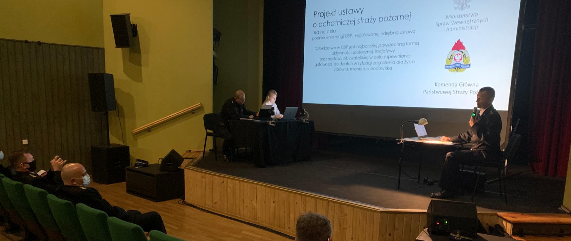 Na scenie Domu Kultury ustawione są dwa stoły. Przy jednym (po lewej) siedzi zastępca Opolskiego Komendanta Wojewódzkiego PSP w Opolu st.bryg. Arkadiusz Kuśmierski umawiający ustawę. Przy drugim stoliku siedzą dwie osoby protokołujące spotkanie. W tle na ekranie wyświetlony slajd prezentacji dotyczącej projektu ustawy. W lewym dolnym roku na krzesłach typu kinowego siedzą uczestnicy konsultacji.