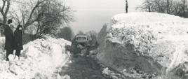 Pług dwustronny na ciężarówce Studebaker odśnieża drogę z błota pośniegowego. Po prawej stronie drogi z góry śniegu sięgającej dwukrotnie ponad kabinę ciężarówki akcję obserwuje człowiek.