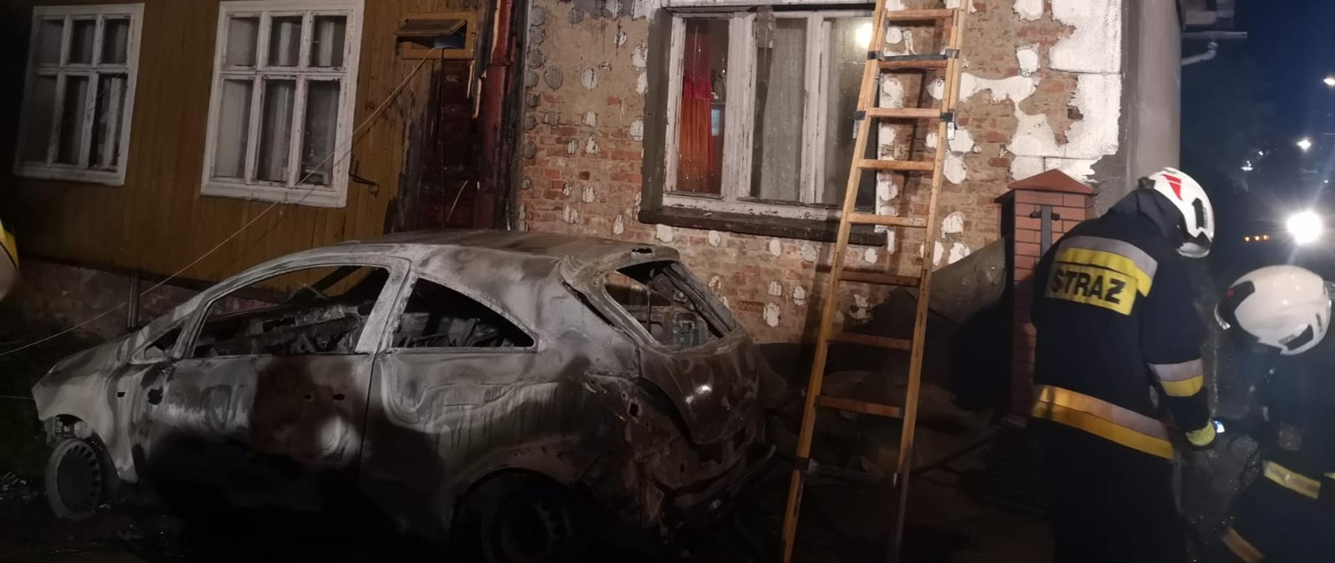 Zdjęcie wykonane w porze nocnej przedstawia budynek mieszkalny ze spaloną elewacją oraz stojący przed nim wrak samochodu osobowego. Z prawej strony strażacy regulują sprzęt oświetleniowy. Na dachu jeden ze strażaków sprawdza czy nie występują gdzieś niedogaszone zarzewia ognia. O budynek oparta jest drabina.