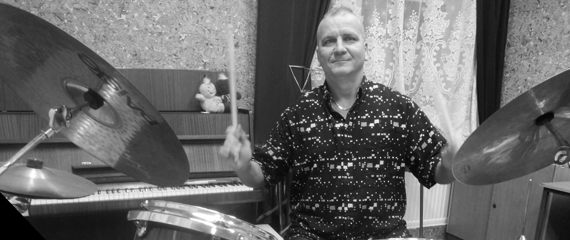 Czarno-białe zdjęcie przedstawiające nauczyciela klasy perkusji - Krzysztofa Palmowskiego podczas gry na perkusji w sali lekcyjnej szkoły.