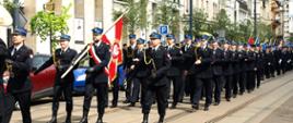 Z lewej Dowódca Kompanii Honorowej i Poczet sztandarowy przechodzący przez ulicę Gdańską w Bydgoszczy, W tle pozostali członkowie Kompanii Honorowej, strażacy w granatowych mundurach wyjściowych. W tle elewacje kremowe budynków przebiegających wzdłuż ul. Gdańskiej.