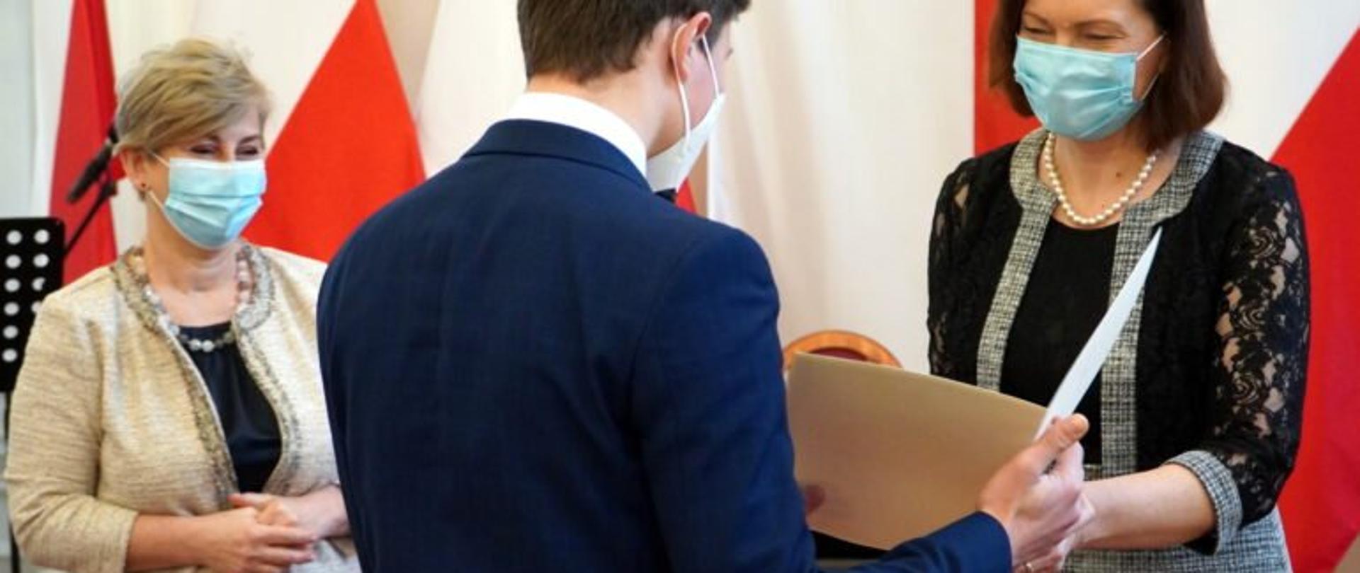 Wojewoda podkarpacki Ewa Leniart wręcza dyplom jednemu z uczniów podczas uroczystości w sali kolumnowej Podkarpackiego Urzędu Wojewódzkiego w Rzeszowie. Po lewej stronie stoi podkarpacki kurator oświaty Małgorzata Rauch. 