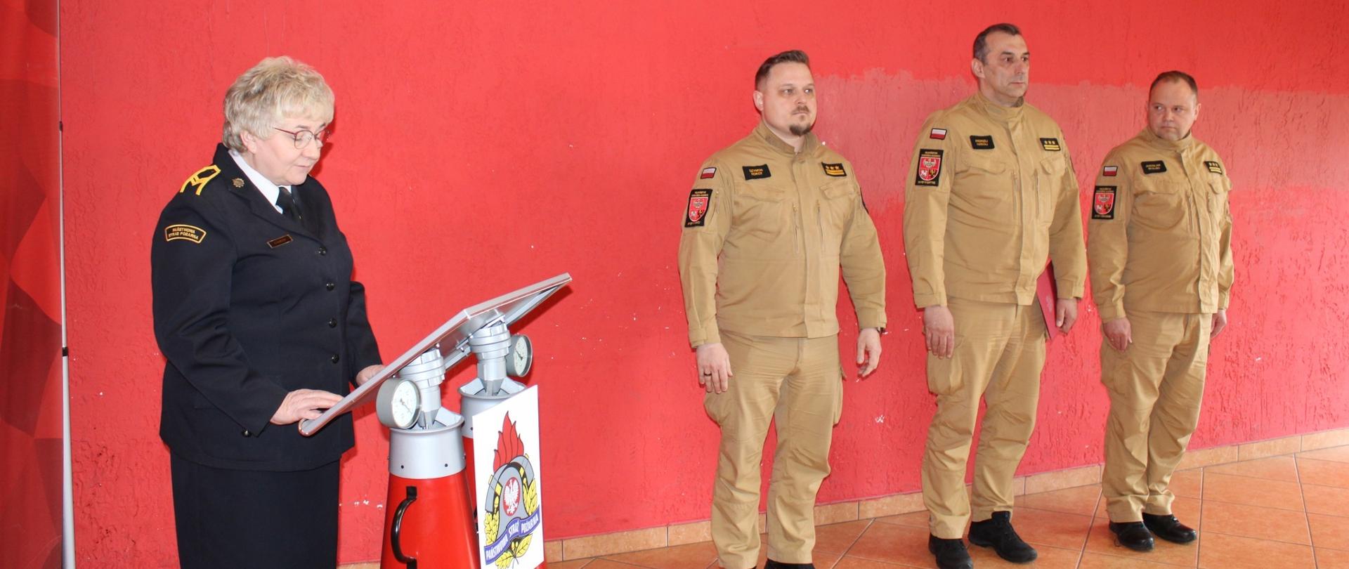 Na tle czerwonej ściany stoi trzech strażaków w jasnych mundurach. Z lewej strony przy mównicy funkcjonariuszka w mundurze wyjściowym.
