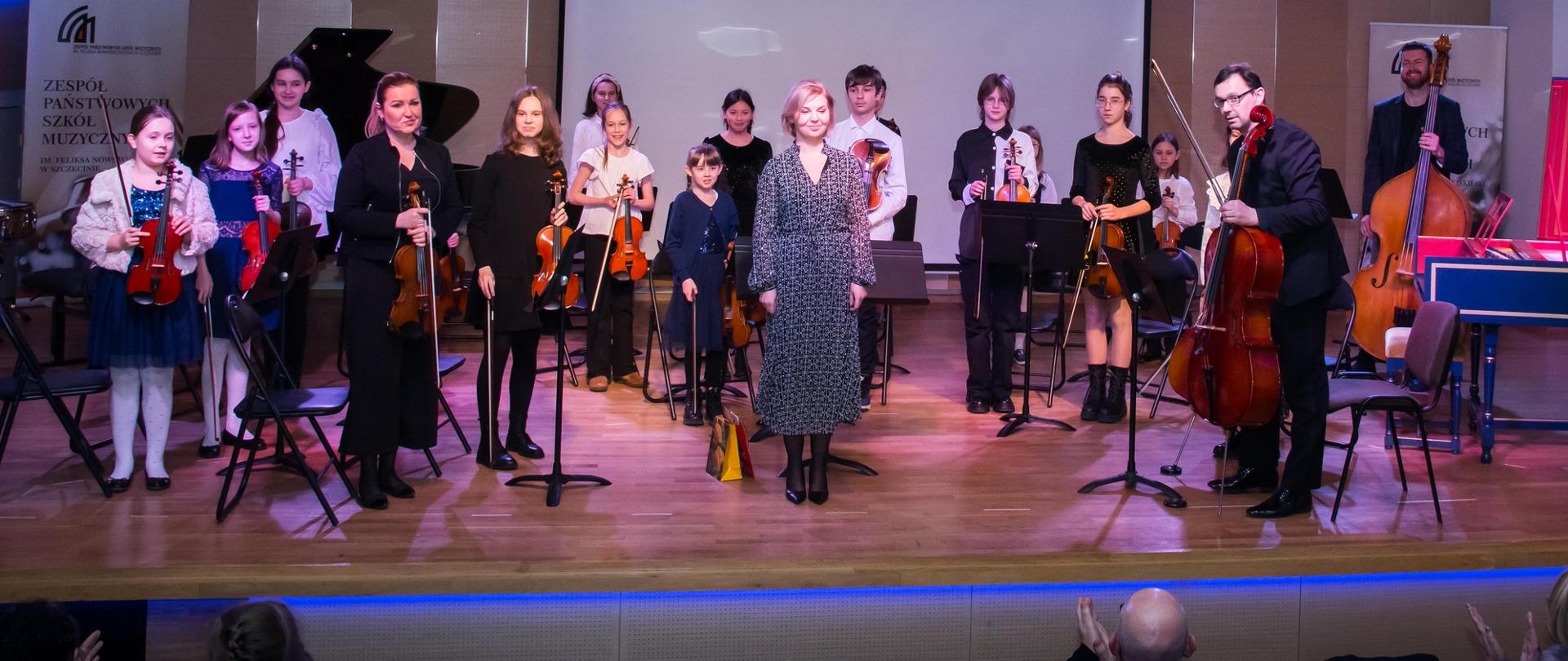 Zdjęcie przedstawia smyczkową orkiestrę kameralną szkoły pierwszego stopnia wraz z nauczycielami na scenie sali koncertowej przed wykonaniem utworu
