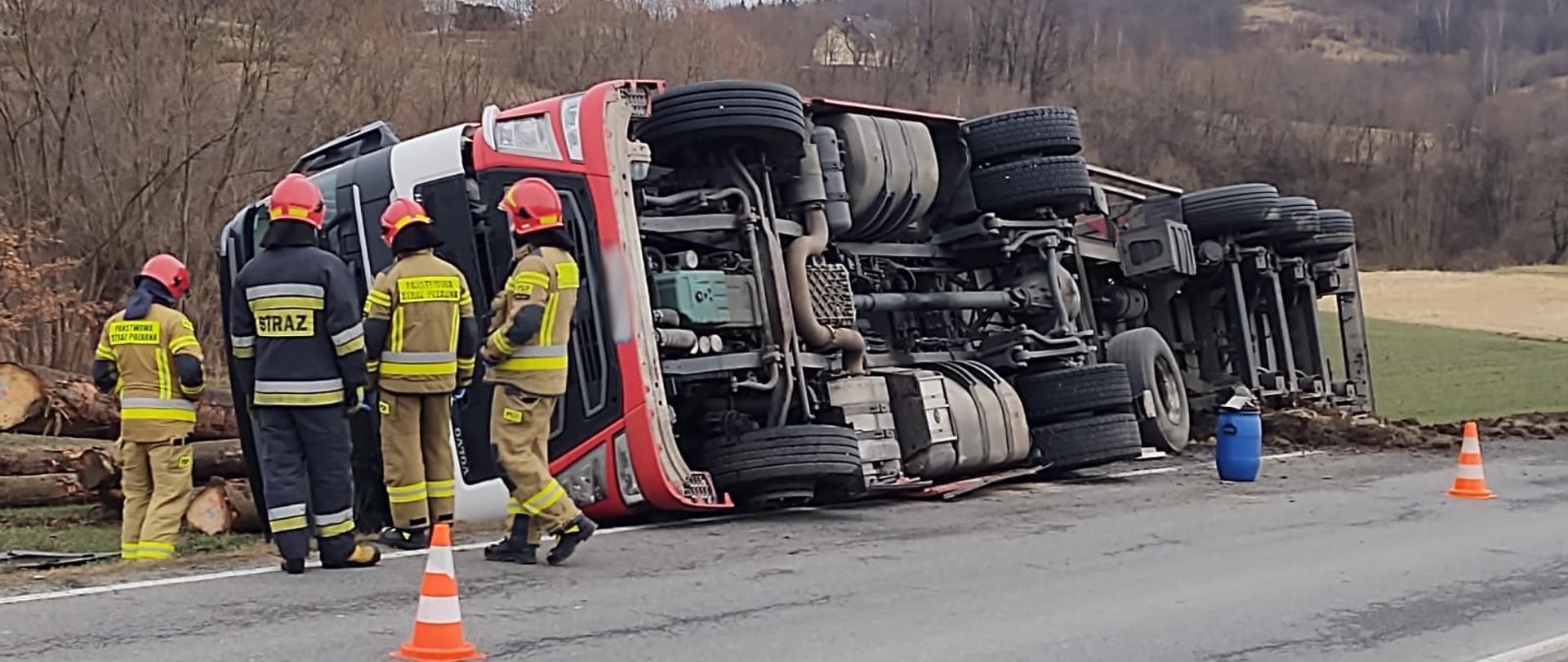 Samochód ciężarowy na boku a obok niego strażacy.