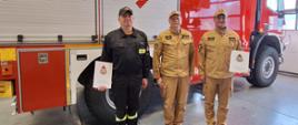 Trzech strażaków stoi przed czerwonym wozem strażackim w garażu jednostki. Dwóch z nich ubranych jest w jasnobrązowe mundury galowe, jeden w czarny mundur koszarowy. Dwie osoby trzymają dyplomy, co sugeruje, że uczestniczą w ceremonii wręczenia wyróżnień lub podziękowań. Za nimi widoczny jest oznaczony numerami pojazd pożarniczy z symbolem ochrony przeciwpożarowej.