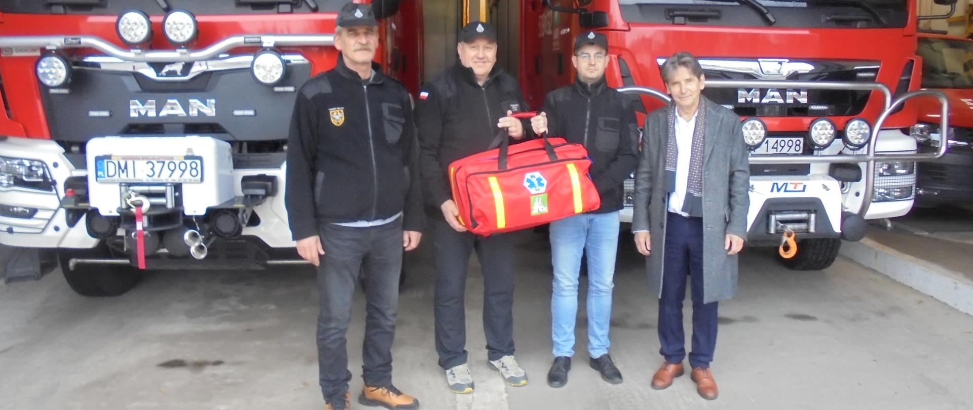 Czterech mężczyzn stoi przed garażem, w którym stoją dwa wozy bojowe straży pożarnej. Dwóch mężczyzn w bluzach OSP trzyma czerwoną torbę z zestawem ratownictwa. obok stoi mężczyzna w cywilnym ubraniu.