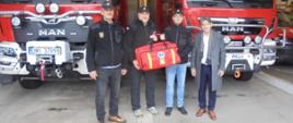 Czterech mężczyzn stoi przed garażem, w którym stoją dwa wozy bojowe straży pożarnej. Dwóch mężczyzn w bluzach OSP trzyma czerwoną torbę z zestawem ratownictwa. obok stoi mężczyzna w cywilnym ubraniu.