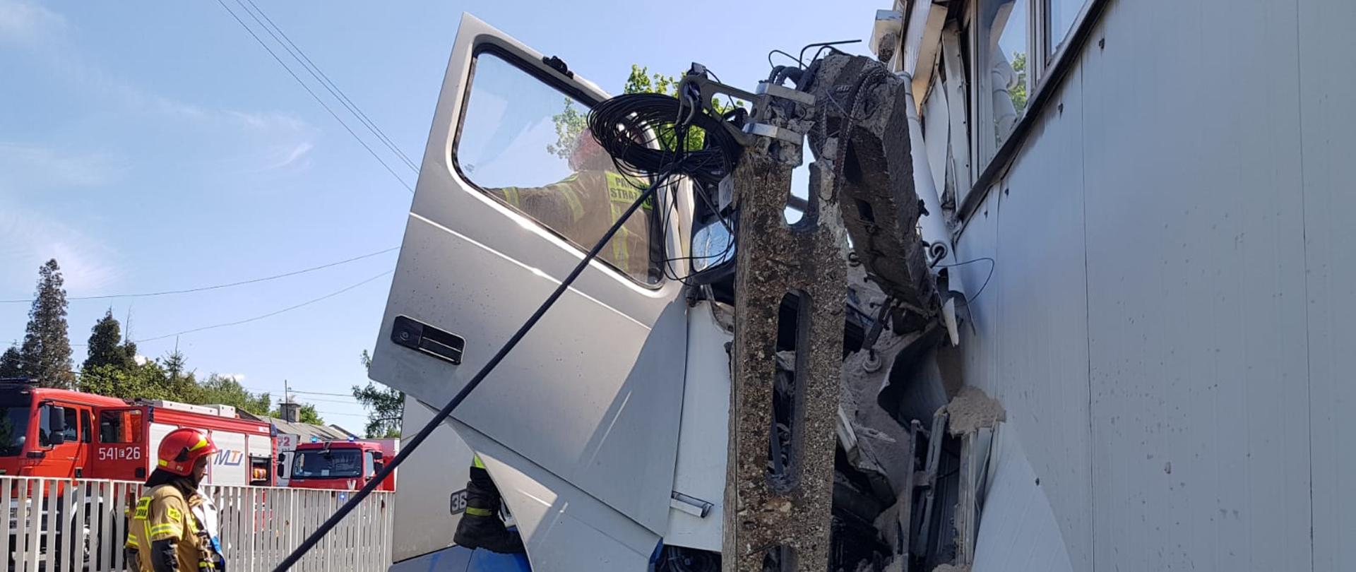 Samochód ciężarowy wbity w ścianę budynku. Przód samochodu jest zgnieciony. Przed samochodem stoi złamany betonowy słup. Po lewej stronie jasne ogrodzenie. Przed ogrodzenie stoi strażak. za ogrodzeniem stoją wozy strażackie. 