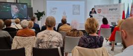 Sala konferencyjna, na ścianie obraz z rzutnika prezentujące przykładowe ekslibrisy, po prawej stronie na ściance z Logo państwowe biblioteki Publicznej przy mównicy kobieta w ciemnym ubraniu. Po lewej stronie telewizor na ekranie którego widać ptaszyna Wróblewskiego – znanego muzyka jazz owego. Na pierwszym planie widać uczestników konkurencji siedzących na krzesłach tyłem do obserwatora
