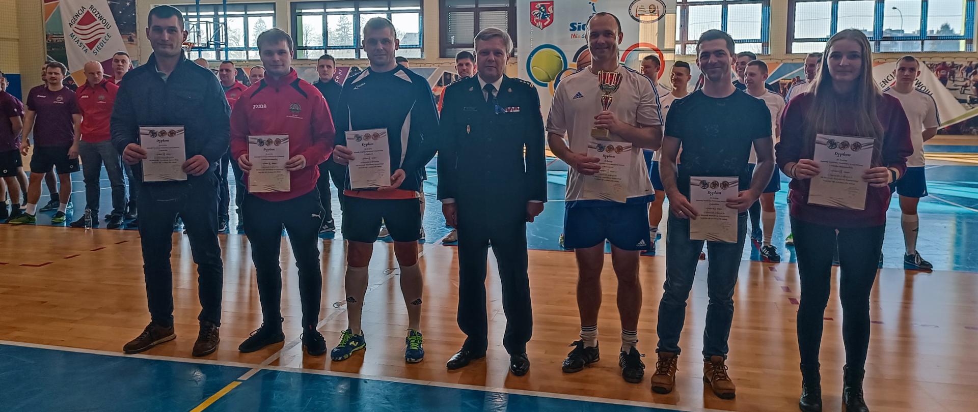 Eliminacje strefowe do mistrzostw strażaków w halowej piłce nożnej. Zdjęcie grupowe kierowników drużyn ze starszym brygadierem Andrzejem Celińskim. Zawodnicy trzymają dyplomy za udział w zawodach.