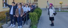Na zdjęciu grupa uczniów, którzy stoją na podjeździe dla niepełnosprawnych prze żółtym budynkiem szkoły. Obok nich na chodniku z ręką do góry stoi kobieta w szarym płaszczyku, czarnej spódnicy i fioletowym berecie. 