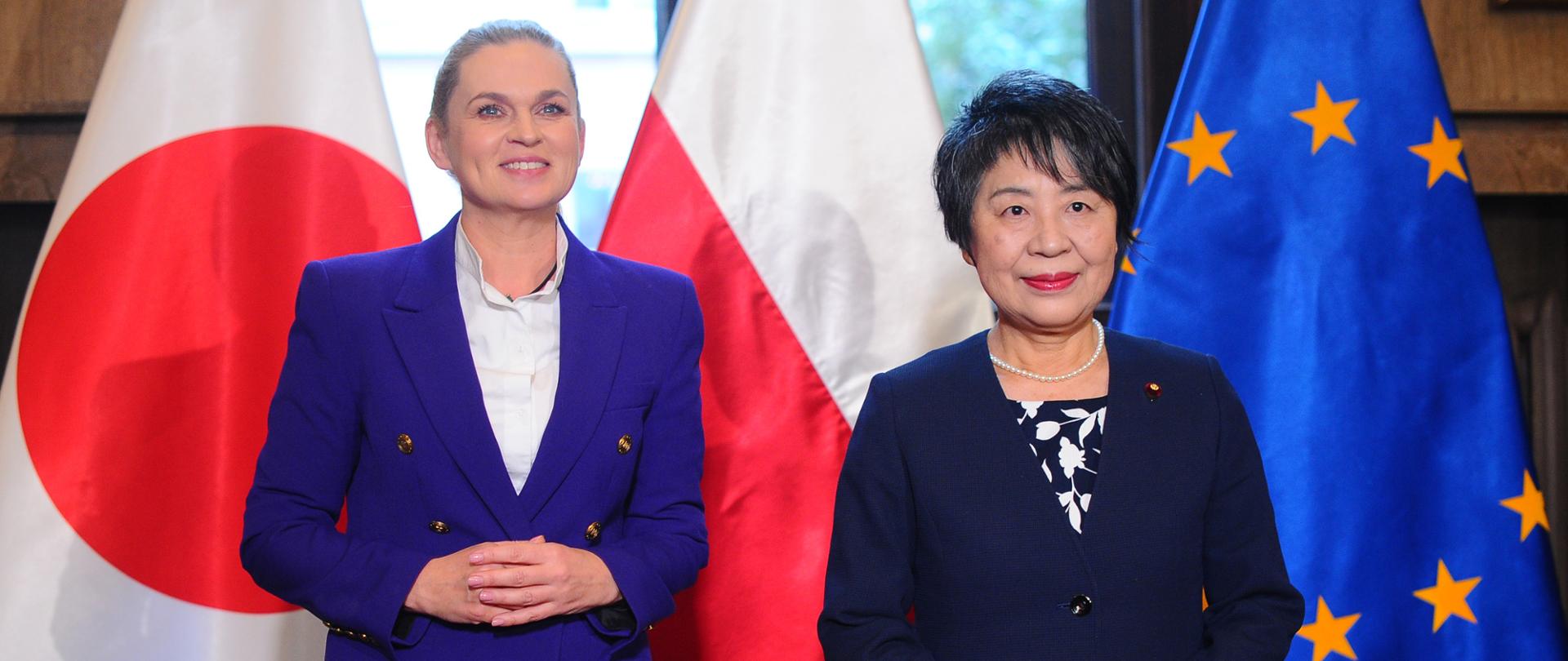 Na zdjęciu, na tle flag Japonii, Polski oraz Unii Europejskiej Stoi po lewej stronie minister edukacji Barbara Nowacka ubrana w białą bluzkę i niebieską marynarkę. Po prawej stronie stoi Minister Spraw Zagranicznych Japonii Yōko Kamikawa ubrana w sukienkę granatową w białe kwiaty i granatową marynarkę. 