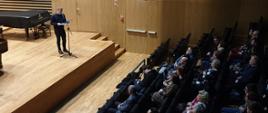Nauczyciel stoi przy mikrofonie i zapowiada koncert na scenie sali koncertowej PSM