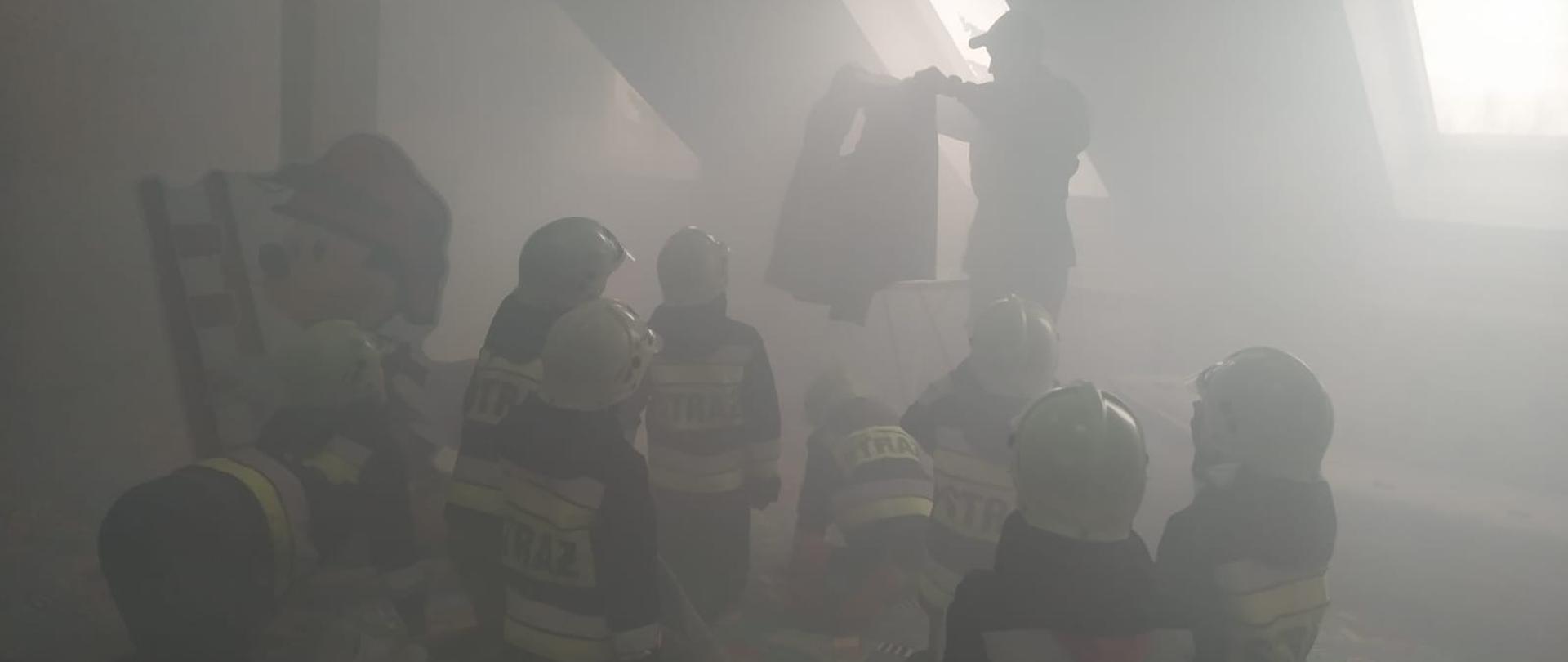 Zdjęcie przedstawia strażaka, który trzyma w rękach koszulę z przepaloną dziurą po prasowaniu. Dzieci trzymają w rękach prądownicę wodną oraz wąż strażacki. Wszyscy znajdują się na sali edukacyjnej Ognik, w której panuje niewielkie zadymienie.