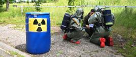 Dwoje strażaków w pełnej odzieży ochronnej klęczy obok niebieskiej beczki oznaczonej symbolem promieniowania, podczas gdy trzecia osoba w odzieży cywilnej prowadzi z nimi rozmowę na otwartym, trawiastym terenie.
Na pierwszym planie widoczna jest niebieska beczka z ostrzeżeniem o promieniowaniu. Obok niej klęczy dwóch strażaków w zielonych skafandrach ochronnych, wyposażonych w czerwone buty oraz aparaty tlenowe. W pobliżu stoi mężczyzna w ciemnej kurtce, który wydaje się przekazywać instrukcje lub omawiać sytuację.
W tle rozciąga się teren z wysoką zielenią, drzewami oraz białą barierą ochronną na tle częściowo zachmurzonego nieba.
Scena przedstawia poważne i profesjonalne działania w terenie otwartym, związane z potencjalnie niebezpieczną substancją. Atmosfera jest skupiona i wskazuje na sytuację wymagającą ostrożności oraz koordynacji działań.
