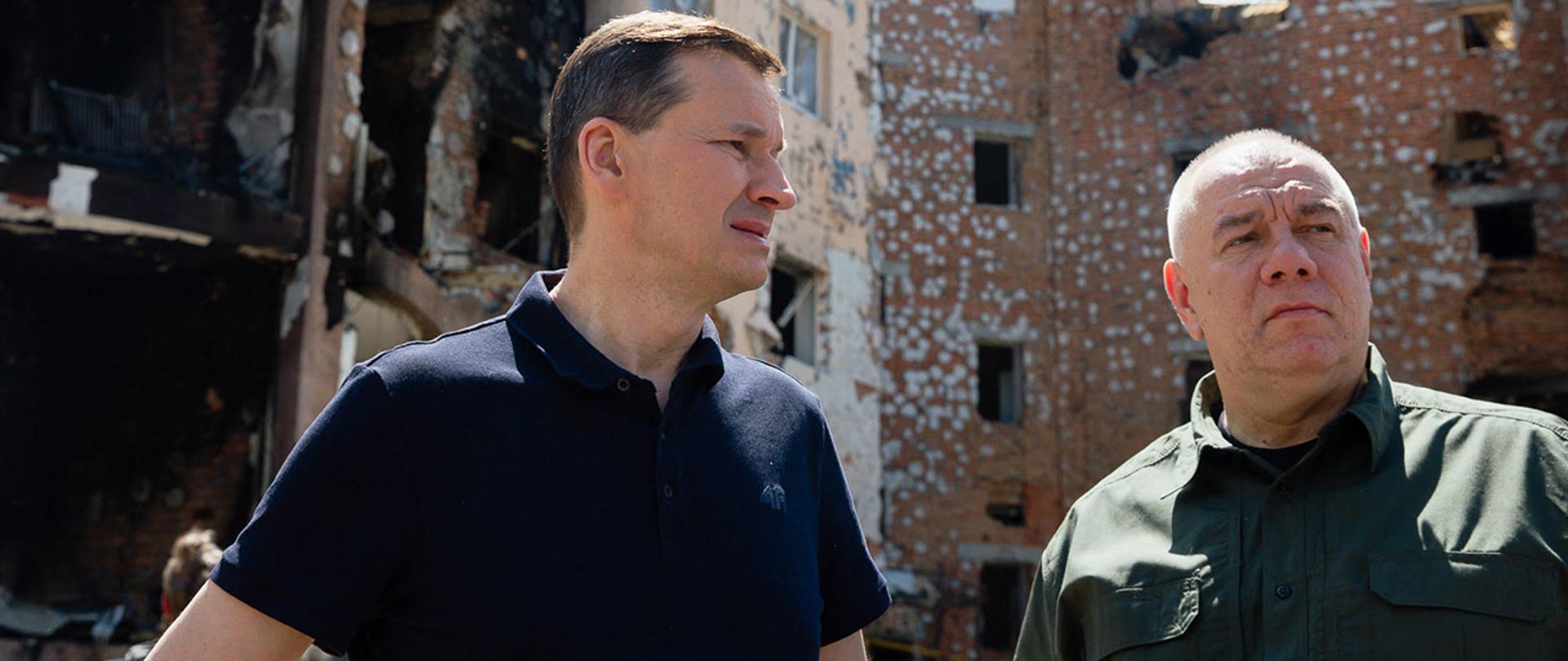 Premier Mateusz Morawiecki i wicepremier Jacek Sasin stoją na tle zniszczonego budynku. 