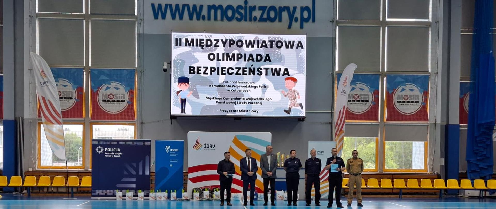 Hala sportowa z niebieską podłogą i żółtymi trybunami po bokach. W centralnej części kadru stoi grupa siedmiu osób ustawionych w linii. Za nimi znajduje się duży ekran cyfrowy, na którym widnieje tytuł wydarzenia: II Międzypowiatowa Olimpiada Bezpieczeństwa.
