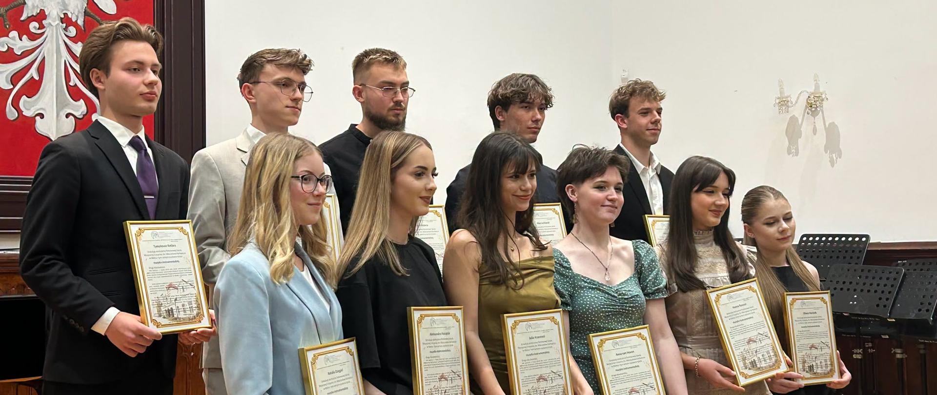 Absolwenci PSM II stopnia trzymający pamiątkowe listy gratulacyjne pozują do zdjęcia grupowego w Sali Królewskiej podczas uroczystości pożegnania dyplomantów, w tle herb Polski.
