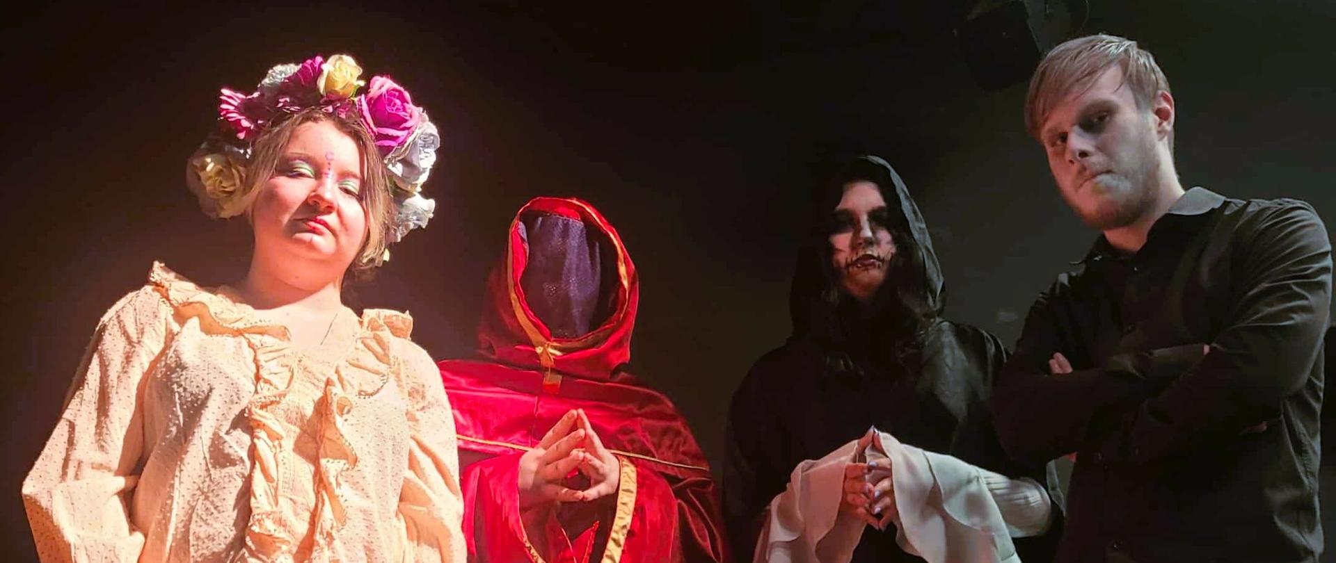 Cztery osoby stoją na scenie w teatralnym, mrocznym oświetleniu. Po lewej stronie znajduje się osoba w jasnej, fantazyjnej sukni i z dużą kwiecistą koroną na głowie. Obok niej stoi postać w czerwonej, zakapturzonej szacie, z twarzą zasłoniętą materiałem. Trzecia osoba ubrana jest w czarny, luźny strój z kapturem i ma charakteryzację przypominającą makijaż grozy. Po prawej stronie stoi osoba w czarnej koszuli, z założonymi rękami, patrząca poważnie przed siebie. Cała czwórka prezentuje teatralne, nieco tajemnicze kreacje.