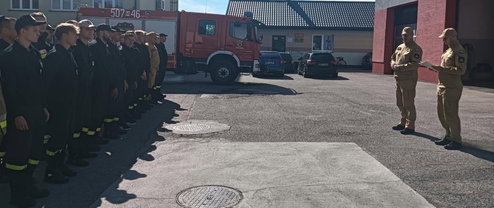 Zdjęcie wykonane na placu przed budynkiem komendy. Z lewej strony w dwóch rzędach stoją strażacy OSP, obok nich stoi wóz strażacki. Z prawej strony widać budynek komendy przed którym stoi Zastępca Komendanta Powiatowego PSP wraz z Dowódcą JRG , który trzyma w rękach zaświadczenia ukończenia szkolenia. 