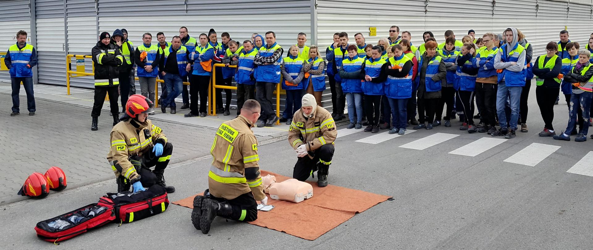 na zdjęciu pracownicy zakładu przyglądają się pokazowi udzielania pierwszej pomocy przez strażaków 