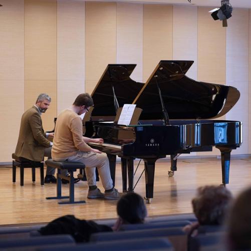 zdjęcie przedstawia ucznia wykonującego ćwiczenie w duecie razem z wykładowcą przy dwóch fortepianach na scenie sali koncertowej