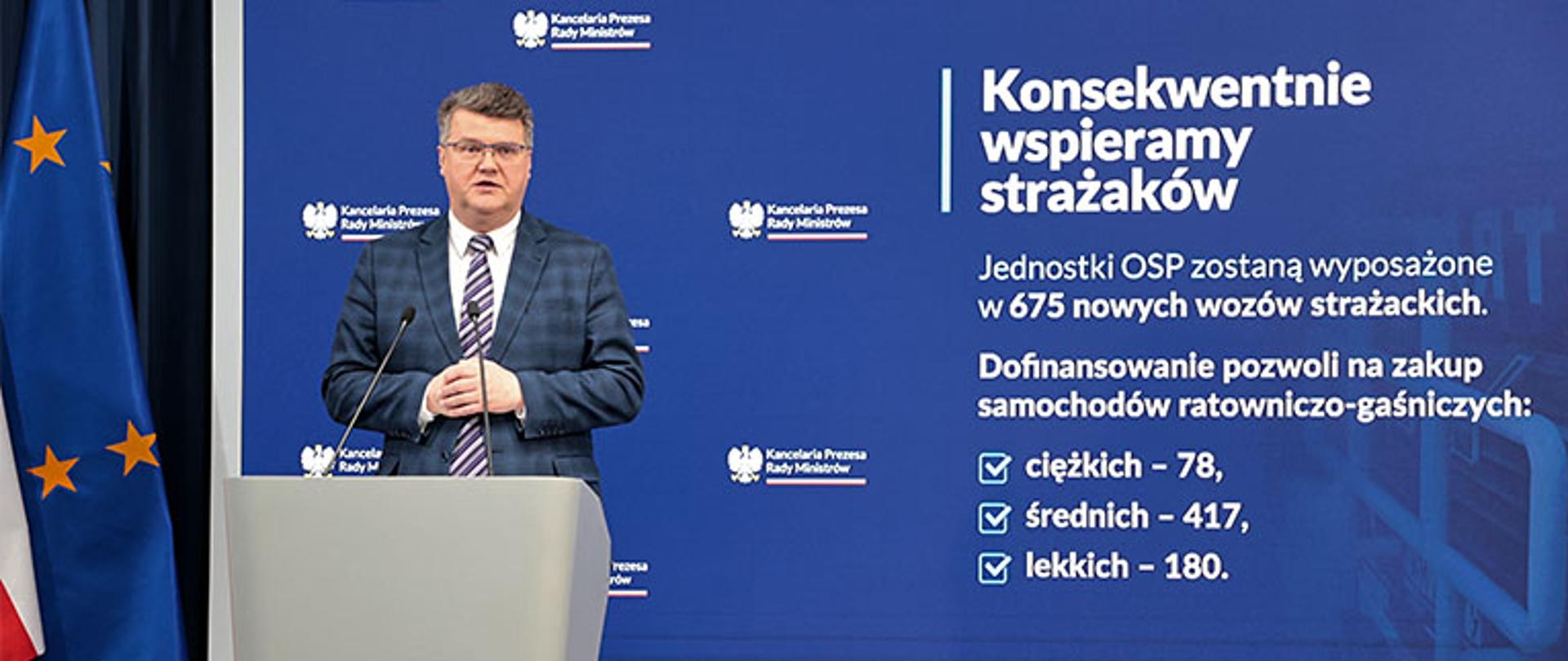 minister Wąsik przemawia z trybuny w tle duży baner z kancelarii prezesa rady ministrów po lewej stronie flaga unii europejskiej