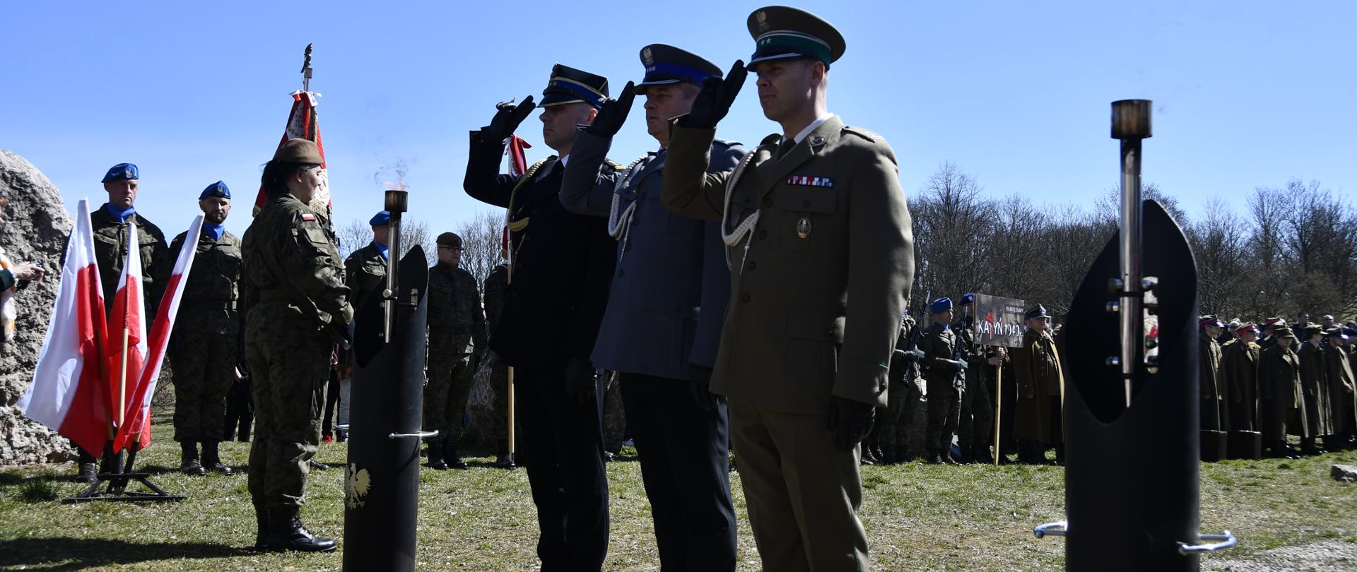 Na zdjęciu widzimy trzech mężczyzn w mundurach trzech służb - Państwowej Straży Pożarnej, Policji, Straży Granicznej - składających wieniec. Mężczyźni salutują.