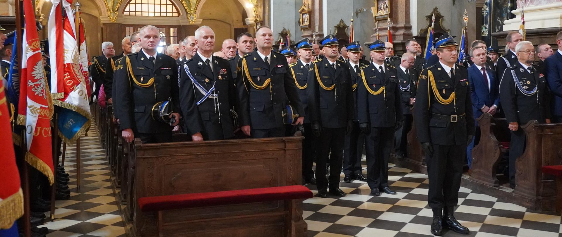 Uczestnicy III Diecezjalnej Pielgrzymki Strażaków w Studziannie. W nawie głównej bazyliki w drewnianych ławkach stoją zaproszeni goście honorowi, w tym Zastępca Łódzkiego Komendanta Wojewódzkiego Państwowej Straży Pożarnej oraz komendanci powiatowi PSP z terenu diecezji. Na środku stoi kompania reprezentacyjna strażaków Państwowej Straży Pożarnej na czele z dowódcą kompanii. Strażacy ubrani są w ciemne mundury reprezentacyjne ze złotymi sznurami, strażacy stojący na baczność w kompani reprezentacyjnej mają na głowach czapki rogatywki, pozostali funkcjonariusze uczestniczący w liturgii trzymają czapki w dłoniach. Po lewej stronie prezentowane są sztandary jednostek. W tle widać rzędy kolejnych ław kościelnych wypełnionych mundurowymi oraz osobami cywilnymi. Wysoko nad nimi rozpościera się zabytkowy, bogato złocony chór, na którym stoją chórzyści. Po prawej stronie marmurowe filary. 