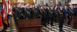 Uczestnicy III Diecezjalnej Pielgrzymki Strażaków w Studziannie. W nawie głównej bazyliki w drewnianych ławkach stoją zaproszeni goście honorowi, w tym Zastępca Łódzkiego Komendanta Wojewódzkiego Państwowej Straży Pożarnej oraz komendanci powiatowi PSP z terenu diecezji. Na środku stoi kompania reprezentacyjna strażaków Państwowej Straży Pożarnej na czele z dowódcą kompanii. Strażacy ubrani są w ciemne mundury reprezentacyjne ze złotymi sznurami, strażacy stojący na baczność w kompani reprezentacyjnej mają na głowach czapki rogatywki, pozostali funkcjonariusze uczestniczący w liturgii trzymają czapki w dłoniach. Po lewej stronie prezentowane są sztandary jednostek. W tle widać rzędy kolejnych ław kościelnych wypełnionych mundurowymi oraz osobami cywilnymi. Wysoko nad nimi rozpościera się zabytkowy, bogato złocony chór, na którym stoją chórzyści. Po prawej stronie marmurowe filary. 