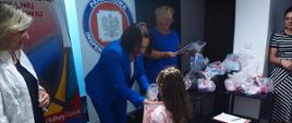 Dziewczynka w różowej sukience otrzymuje prezent od kobiety w niebieskim kostiumie Państwowego Powiatowego Inspektora Sanitarnego w Jarosławiu, a inne kobiety stoją obok, pośród zapakowanych prezentów.