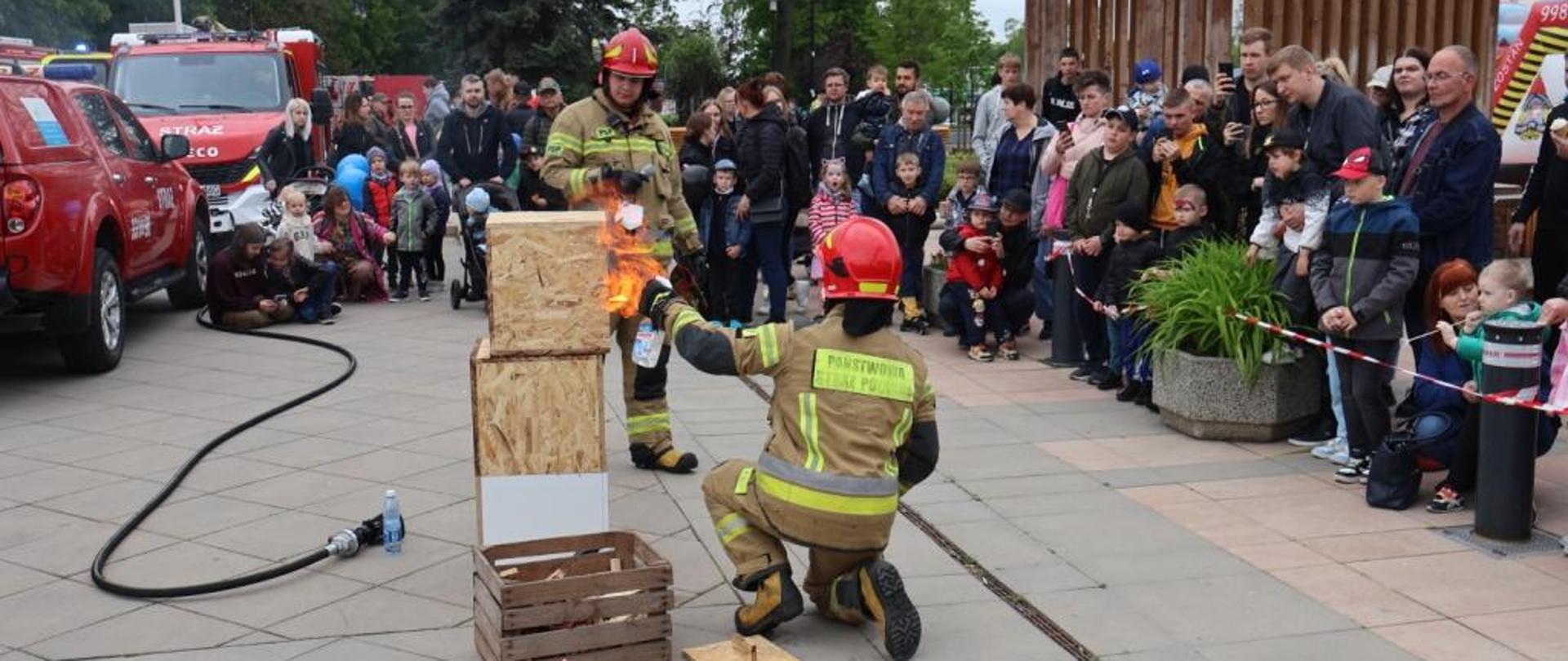 Zdjęcie wykonane na Placu Jagiellońskim w Radomiu podczas rodzinnego pikniku strażackiego zorganizowanego po zakończeniu obchodów powiatowego dnia strażaka. Na zdjęciu widać dwóch strażaków ubranych w hełmy oraz specjalistyczną odzież ochronną. Pomiędzy strażakami znajduje się konstrukcja, która zostanie podpalona w formie pokazu. Dookoła strażaków znajdują się ludzie, osoby odwiedzające piknik obserwujące pokaz. 