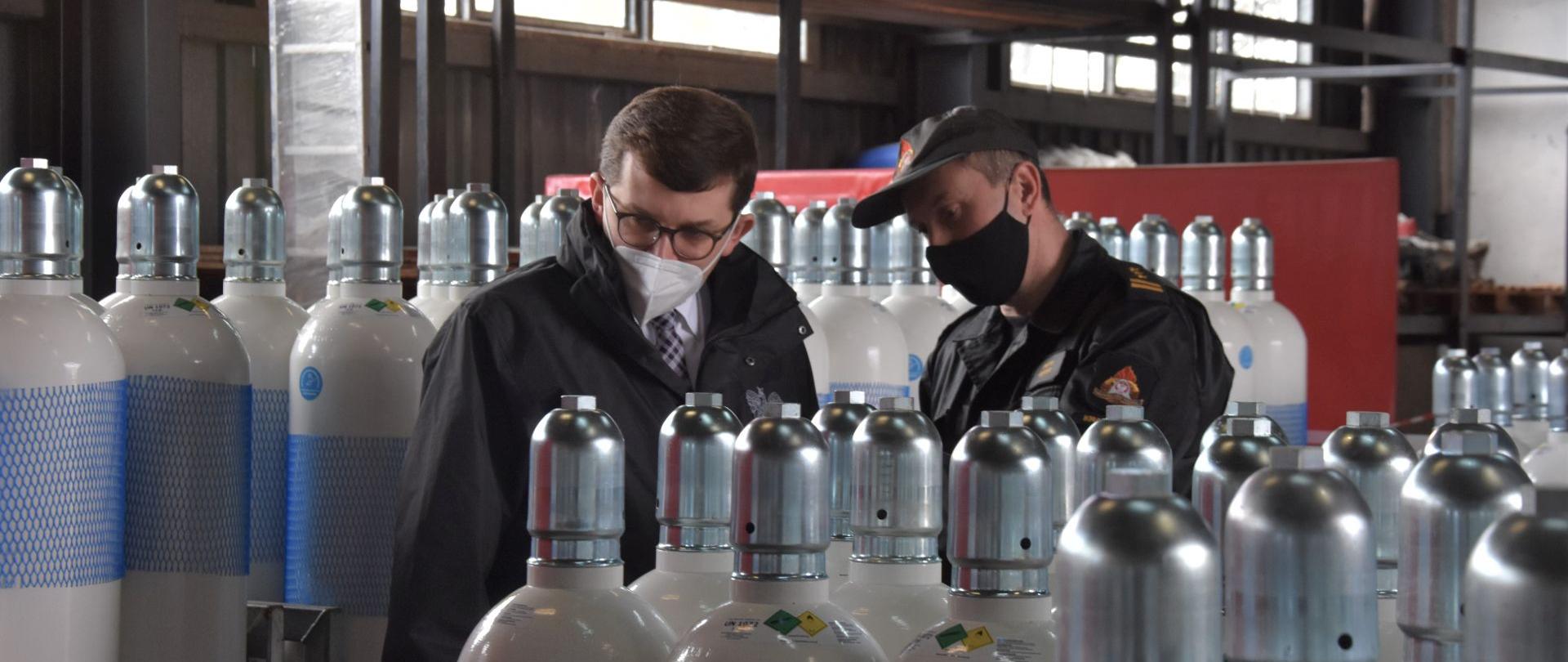 Wojewoda Małopolski wraz z Małopolskim Komendantem Wojewódzkim PSP wizytują bank tlenu. Stoją wewnątrz magazynu. Wokół bardzo dużo butli z tlenem. 