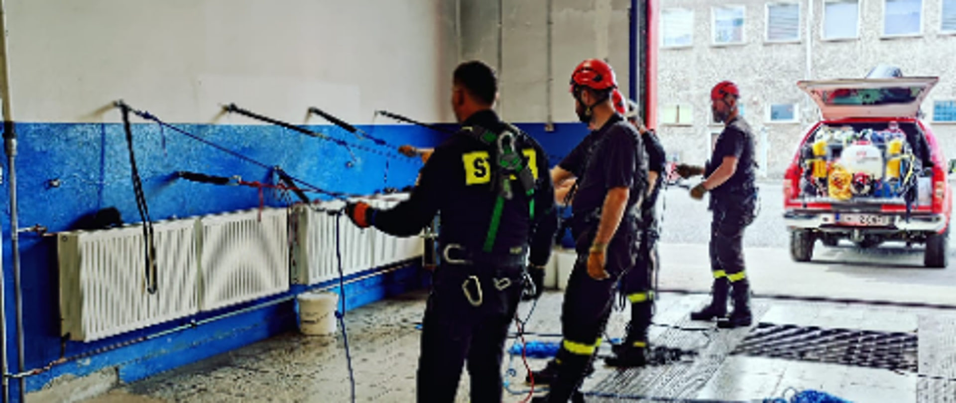 Na zdjęciu znajdują się ratownicy podczas ćwiczenia z ratownictwa wysokościowego, tj. 4 strażaków podpiętych do lin zamocowanych w uchwytach na ścianie nw garażu, w tle stoi samochód z otwartą klapą bagażnika