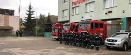 Poczet flagowy podnosi flagę na maszt strażacy stoją przed samochodami gaśniczymi w rzędzie oddając honor.