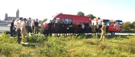 Ćwiczenia z udziałem SGRWN. Zdjęcie przedstawia teren nad zbiornikiem retencyjnym w Jutrosinie.Na tle wozu strażackiego typu bus, do którego podczepiona jest przyczepa z łodzią stoją uczestnicy ćwiczeń - strażacy OSP i PSP. Na przeciwko nich w mundurze koloru piaskowego stoi strażak PSP, który przemawia do zebranych. Z prawej strony widać fragmenty dwóch wozów bojowych, a w oddali widoczny jest kościół w Jutrosinie.