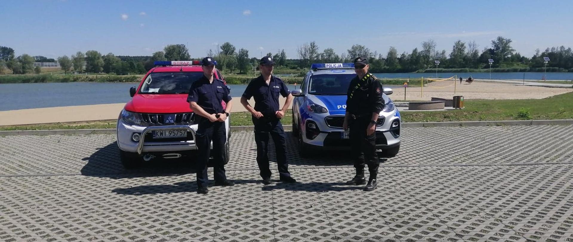 Na zdjęciu dwóch funkcjonariuszy Policji oraz jeden funkcjonariusz Państwowej Straży Pożarnej. Za nimi radiowóz Policji oraz Straży Pożarnej na tle akwenu.