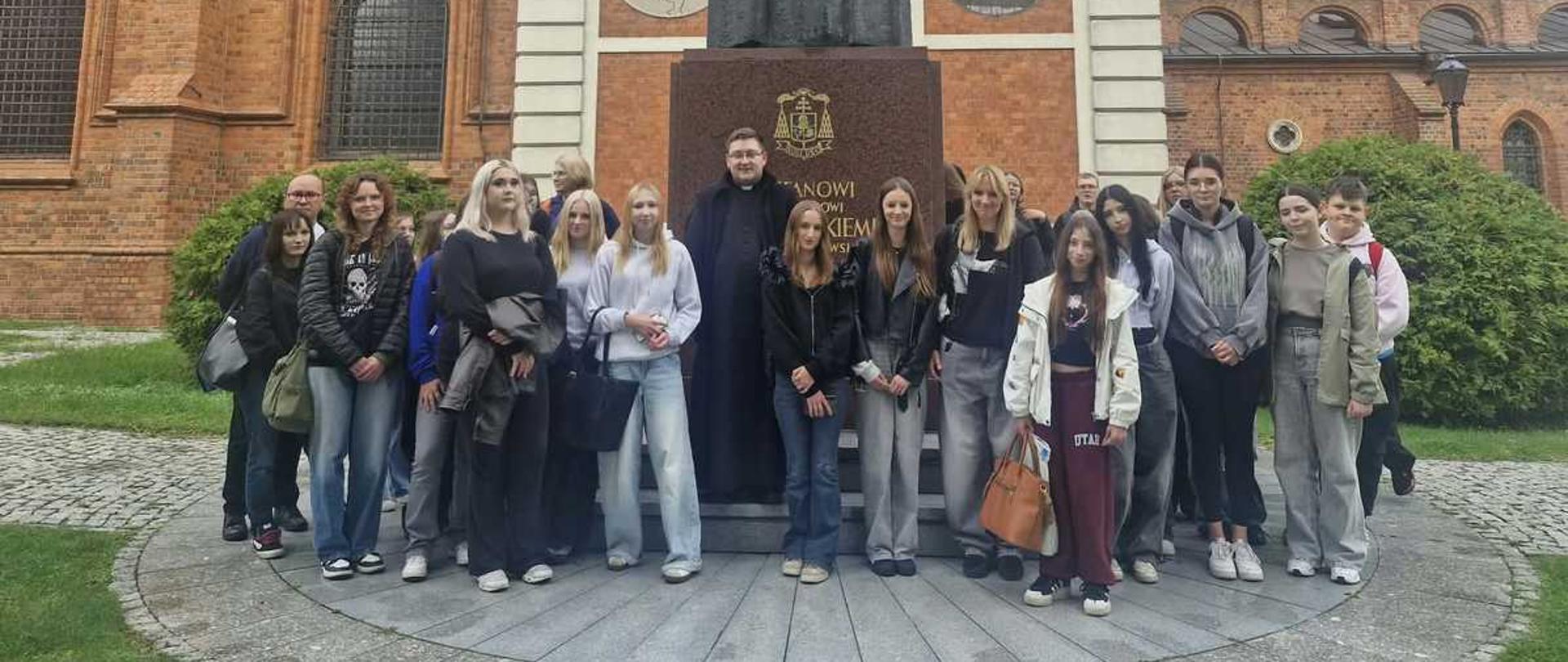 "Zdjęcie przedstawia dużą grupę młodzieży szkolnej oraz księdza, którzy pozują do wspólnego zdjęcia na tle monumentalnego pomnika i zabytkowego, ceglanego kościoła.
W centrum dolnej części kadru stoi grupa około dwudziestu osób, składająca się głównie z nastoletnich dziewcząt i chłopców w codziennych ubraniach (bluzy, jeansy, sneakersy, kurtki). Dokładnie w środku grupy stoi ksiądz ubrany w długą, czarną sutannę i białą koloratkę pod szyją. Wszyscy są zwróceni przodem do obiektywu i stoją na półkolistym podwyższeniu wyłożonym jasną kostką brukową.
Detale drugiego planu (pomnik):
• Bezpośrednio za grupą wznosi się wysoki pomnik kardynała Stefana Wyszyńskiego.
• Postać duchownego, wykonana z ciemnego materiału, stoi na potężnym, brązowo-czerwonym, granitowym postumencie z wyrytym napisem oraz herbem. Kardynał przedstawiony jest w pełnym stroju liturgicznym z piuską na głowie.
• Za pomnikiem znajduje się jasna, architektoniczna ścianka ozdobiona kolumnami, prostokątnymi tablicami z napisami oraz dwoma okrągłymi medalionami (płaskorzeźbami).
Detale otoczenia i tła:
• Architektura: Całość kompozycji domyka w tle ogromny, zabytkowy kościół z czerwonej cegły (prawdopodobnie bazylika katedralna we Włocławku). Po lewej i prawej stronie widoczne są strzeliste, gotyckie okna oraz zdobienia murów.
• Otoczenie: Po bokach placu z pomnikiem rosną zielone, gęste krzewy i trawniki. Pogoda jest pochmurna, a światło dzienne rozproszone.
Kadrowanie zdjęcia jest poziome, ujęcie wykonano na wprost z odległości kilkunastu metrów, aby w pełni ukazać zarówno zgromadzonych ludzi, jak i monumentalną architekturę za nimi."