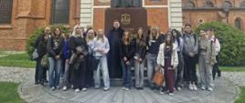 "Zdjęcie przedstawia dużą grupę młodzieży szkolnej oraz księdza, którzy pozują do wspólnego zdjęcia na tle monumentalnego pomnika i zabytkowego, ceglanego kościoła.
W centrum dolnej części kadru stoi grupa około dwudziestu osób, składająca się głównie z nastoletnich dziewcząt i chłopców w codziennych ubraniach (bluzy, jeansy, sneakersy, kurtki). Dokładnie w środku grupy stoi ksiądz ubrany w długą, czarną sutannę i białą koloratkę pod szyją. Wszyscy są zwróceni przodem do obiektywu i stoją na półkolistym podwyższeniu wyłożonym jasną kostką brukową.
Detale drugiego planu (pomnik):
• Bezpośrednio za grupą wznosi się wysoki pomnik kardynała Stefana Wyszyńskiego.
• Postać duchownego, wykonana z ciemnego materiału, stoi na potężnym, brązowo-czerwonym, granitowym postumencie z wyrytym napisem oraz herbem. Kardynał przedstawiony jest w pełnym stroju liturgicznym z piuską na głowie.
• Za pomnikiem znajduje się jasna, architektoniczna ścianka ozdobiona kolumnami, prostokątnymi tablicami z napisami oraz dwoma okrągłymi medalionami (płaskorzeźbami).
Detale otoczenia i tła:
• Architektura: Całość kompozycji domyka w tle ogromny, zabytkowy kościół z czerwonej cegły (prawdopodobnie bazylika katedralna we Włocławku). Po lewej i prawej stronie widoczne są strzeliste, gotyckie okna oraz zdobienia murów.
• Otoczenie: Po bokach placu z pomnikiem rosną zielone, gęste krzewy i trawniki. Pogoda jest pochmurna, a światło dzienne rozproszone.
Kadrowanie zdjęcia jest poziome, ujęcie wykonano na wprost z odległości kilkunastu metrów, aby w pełni ukazać zarówno zgromadzonych ludzi, jak i monumentalną architekturę za nimi."