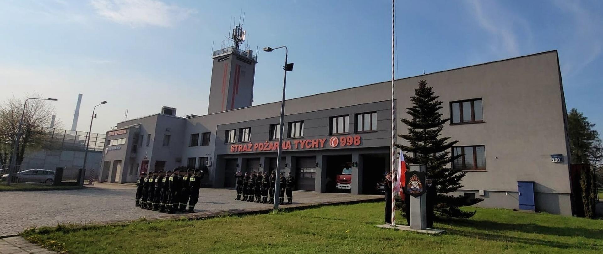 Strażacy podczas uroczystej zmiany służby na której podniesiono flagę na maszt. Strażacy na tle budynku Komendy Miejskiej Państwowej Straży Pożarnej w Tychach