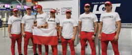 Zdjęcie przedstawia drużyne z polski na lotnisku. Zdjęcie przedstawia 7 osób ubranych na biało z czerwonymi spodniami.
