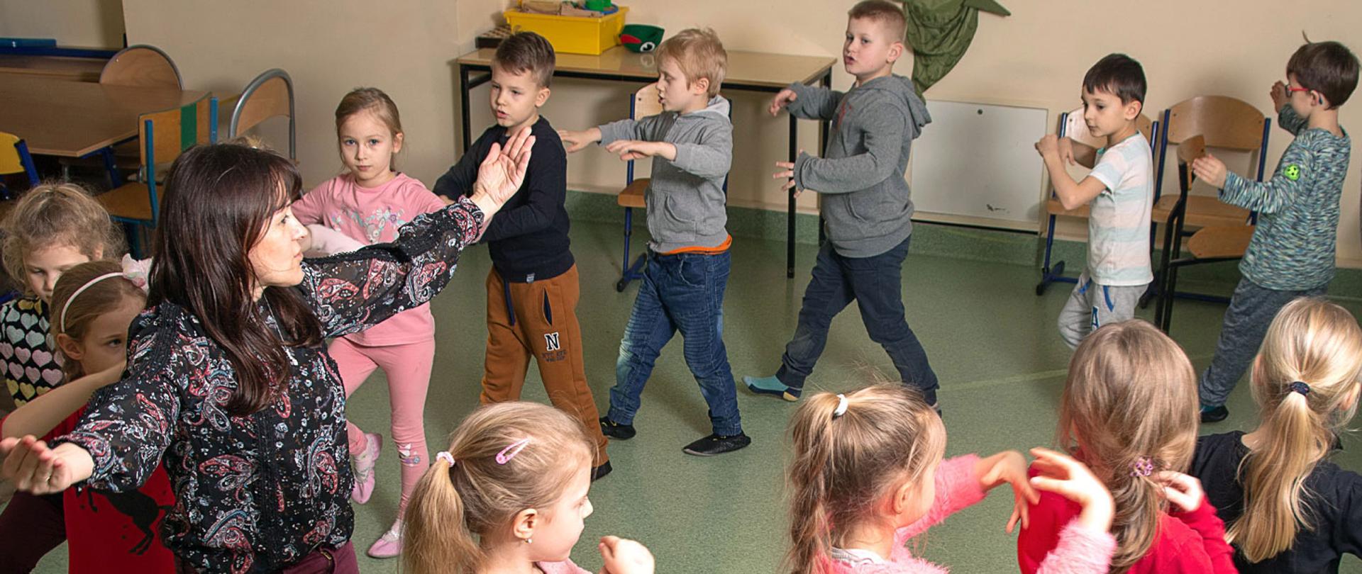 Zdjęcie przedstawia grupę małych dzieci tańczących w kółku wraz ze swoją nauczycielką, wszyscy mają uniesione ręce do góry.