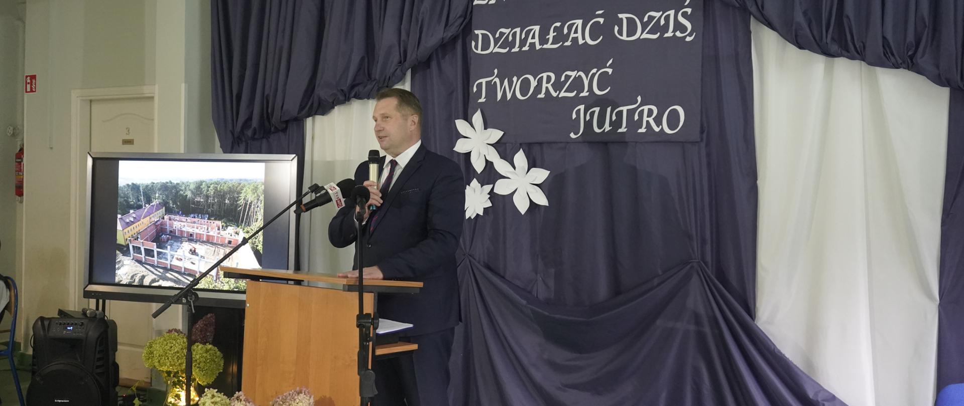 Pod ścianą zasłoniętą czarnym materiałem z napisem Znać wzorce, działać dziś, tworzyć jutro stoi za mównicą minister Czarnek i mówi do dwóch mikrofonów na stojakach.