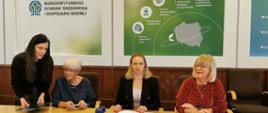 Zdjęcie przedstawia moment podpisania dofinansowania na przedsięwzięcie pod tytułem „Zmniejszenie zużycia energii i emisji CO2 z wykorzystaniem odnawialnych źródeł energii w 6 Zakładach Leczniczych Stowarzyszenia MONAR”. Na zdjęciu widać (zaczynając od lewej strony) koordynator projektu ze strony Narodowego Funduszu Ochrony Środowiska i Gospodarki Wodnej Joanna Jaworek, przewodnicząca zarządu głównego stowarzyszenia MONAR Elżbieta Zielińska, zastępca dyrektora w departamencie transformacji energetycznej w Narodowym Funduszu Ochrony Środowiska i Gospodarki Wodnej Magdalena Bodzenta, sekretarz zarządu głównego stowarzyszenia MONAR Jagoda Władoń- Wilecka. Panie siedzą przy drewnianym stole, na tle loga Narodowego Funduszu Ochrony Środowiska i Gospodarki Wodnej. Na stole leżą teczki z umowami, które przedstawiciele instytucji podpisują.