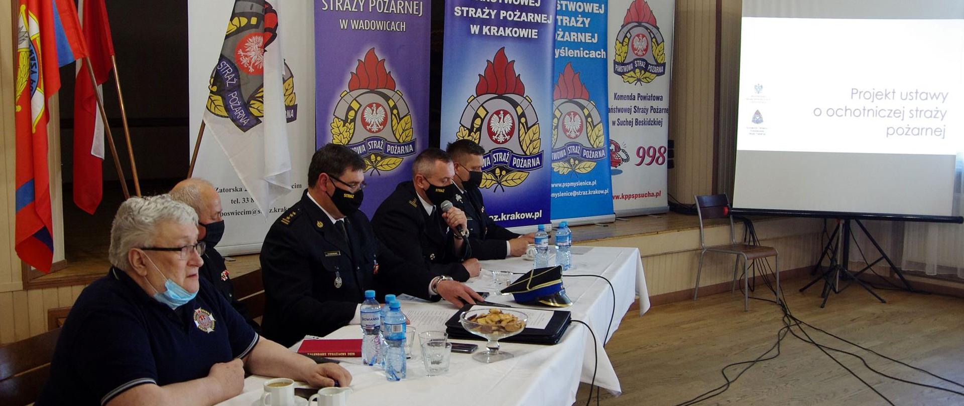Na zdjęciu widzimy strażaków PSP i OSP siedzących przy stole wewnątrz remizy OSP Chocznia. Uczestniczą oni w spotkaniu mającym na celu konsultację zapisów projektu ustawy o OSP. W tle widoczne banery Komendy Wojewódzkiej i Komend Powiatowych, których przedstawiciele uczestniczą w spotkaniu.