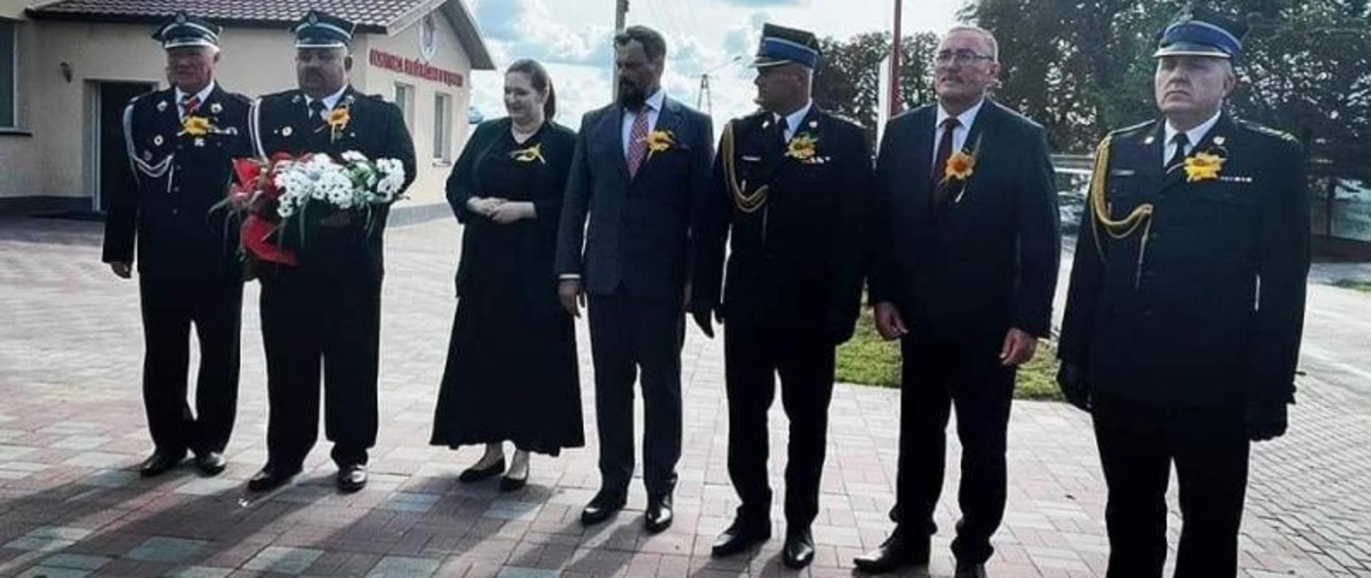 Na zdjęciu stoją strażacy w mundurach galowych oraz trzy osoby cywilne. Stoją oni w szeregu na polbruku.