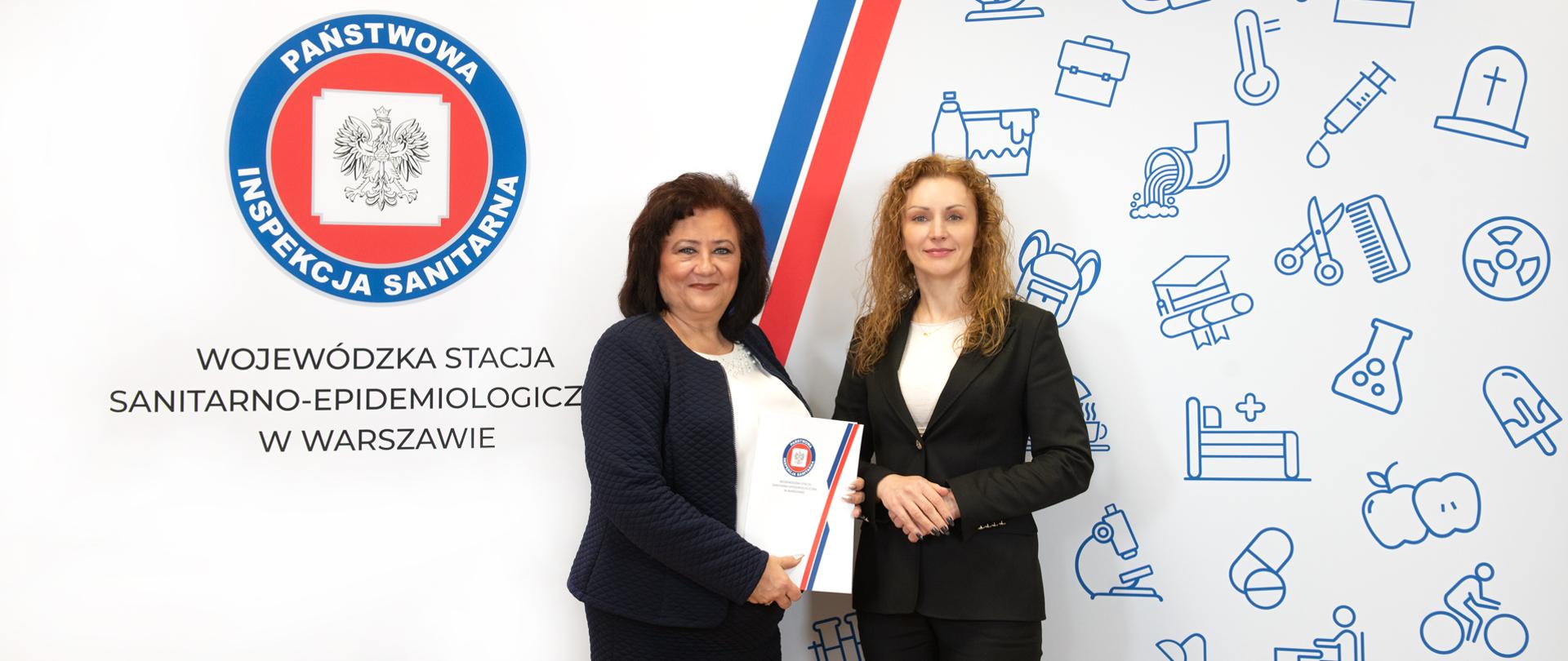 Dwie kobiety w strojach formalnych stoją na tle ścianki z logo Państwowej Inspekcji Sanitarnej. Jedna trzyma w rękach teczkę z dokumentem powołania. 