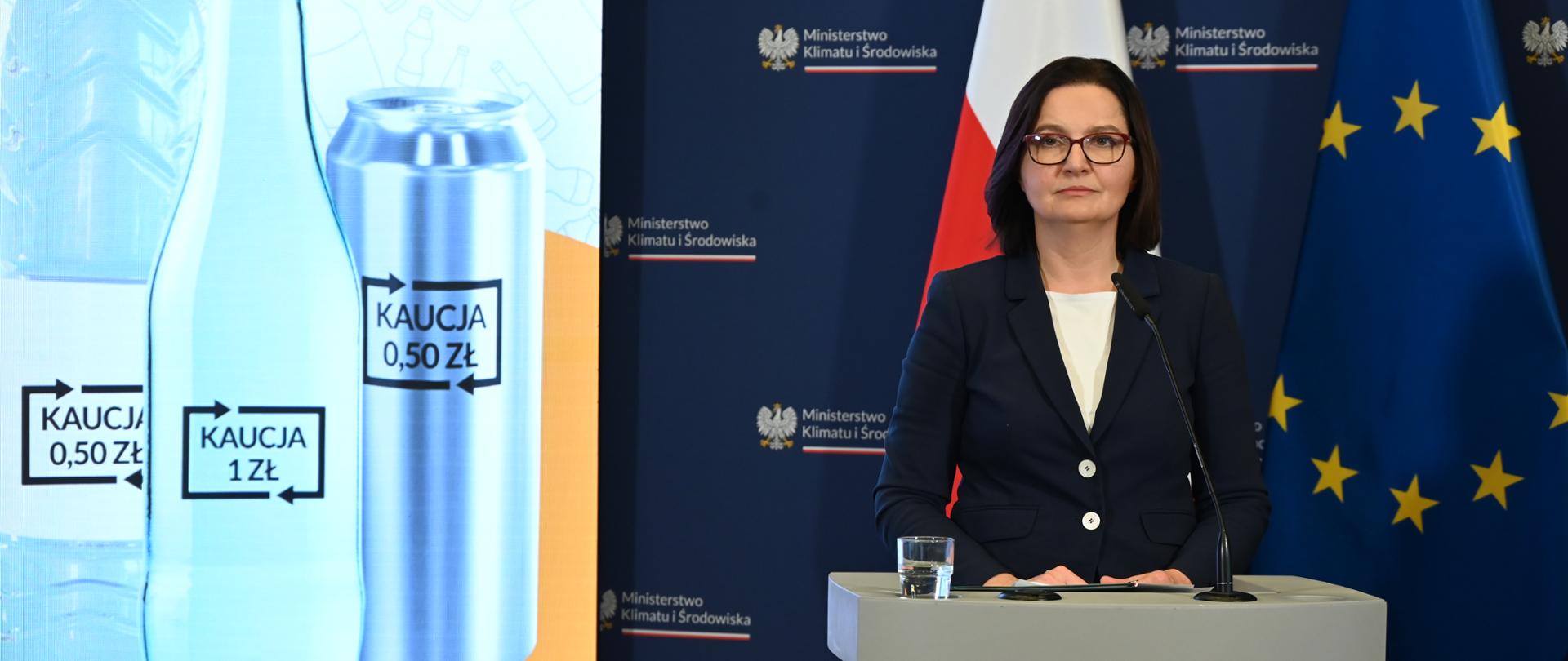 Ministra Anita Sowińska stoi za mównicą. Za nią po lewej stronie fragment telebimu, na którym wyświetla się prezentacja dot. systemu kaucyjnego. Po prawej stronie dwie flagi - flaga Polski i flaga UE. 